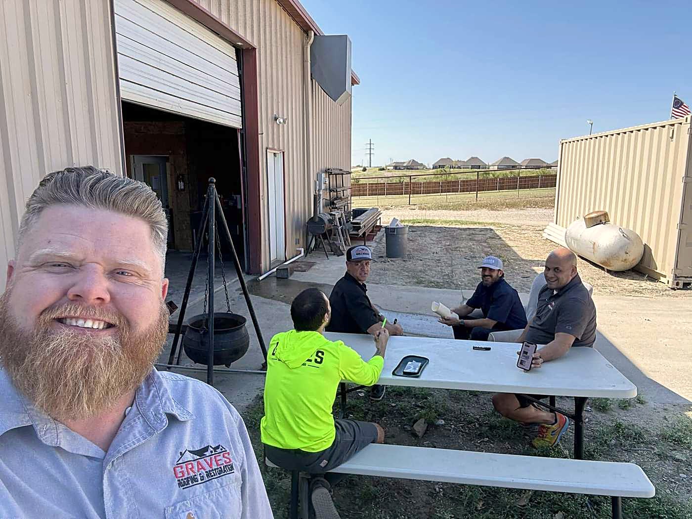 A man in a Graves Roofing shirt in front of a metal building, with four people sitting at tables behind him